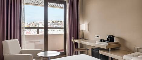 Ein Hotelzimmer mit Bett, Schreibtisch mit Kaffeemaschine, Stuhl, einem kleinen Tisch und Blick auf eine Stadtlandschaft durch große Fenster.