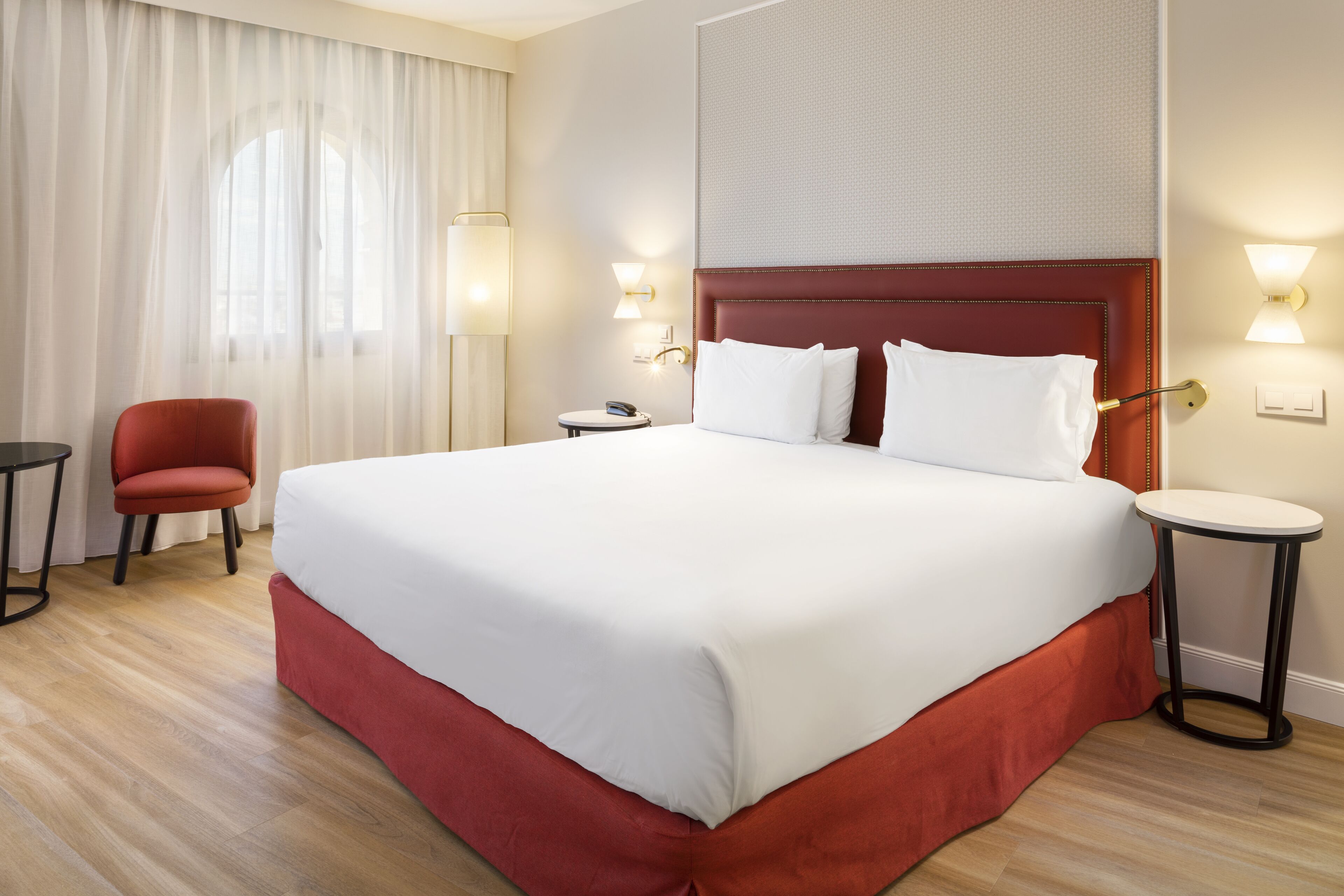 Una habitació d'hotel amb un llit gran, una cadira vermella, una taula rodona petita i una làmpada.