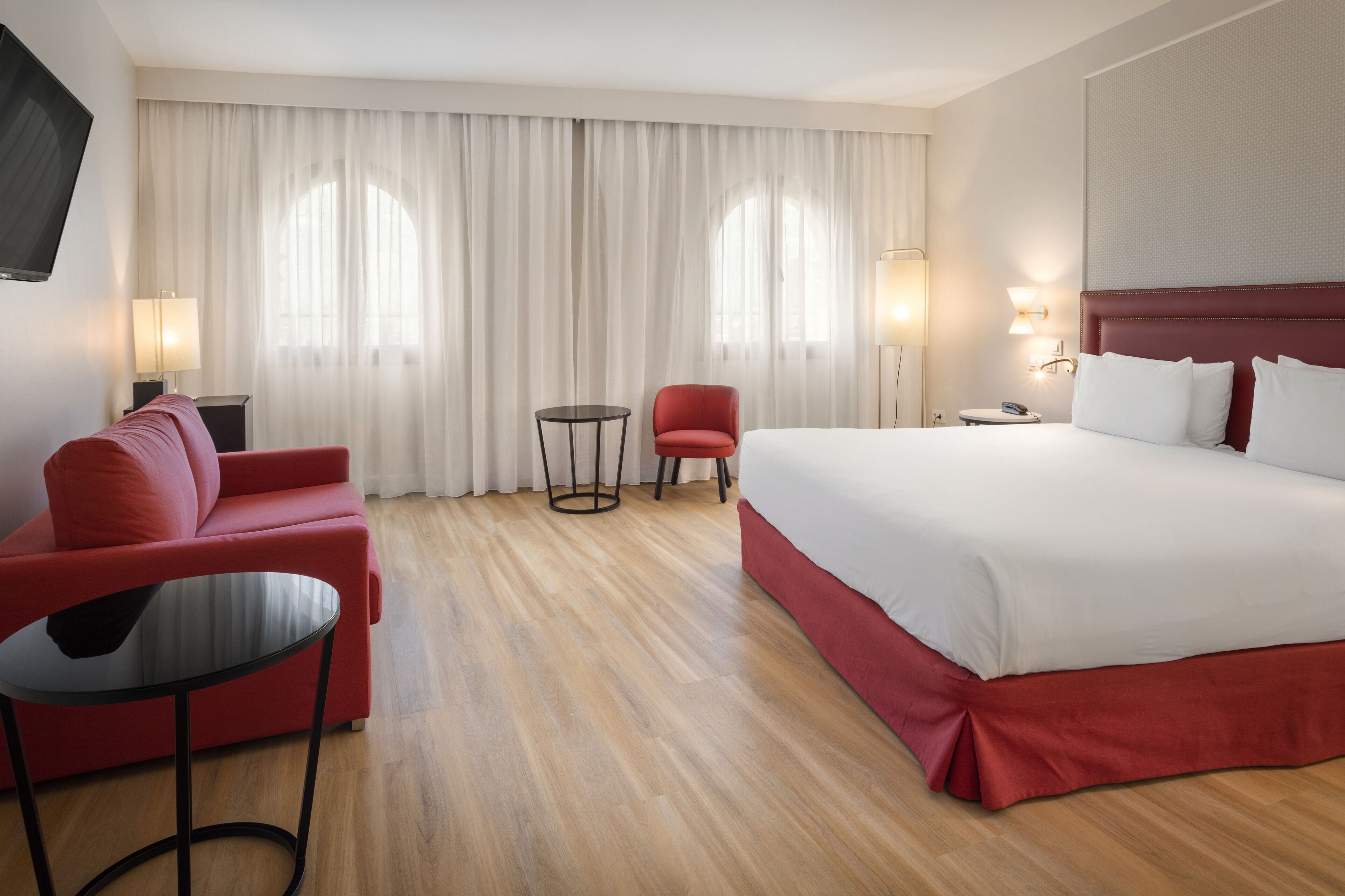 A hotel room with a large bed, a red sofa, a small table, and a TV mounted on the wall.