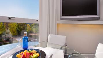 Habitación de hotel con una mesa de comedor preparada para dos personas, un televisor de pantalla plana en la pared y vista a un paisaje verde por la ventana.