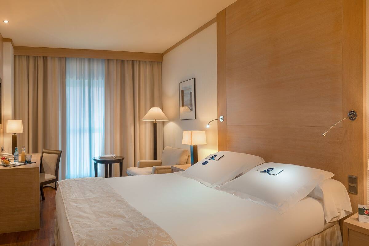 A hotel room with a large bed, a desk with a television, a chair, a small table, and a lamp.
