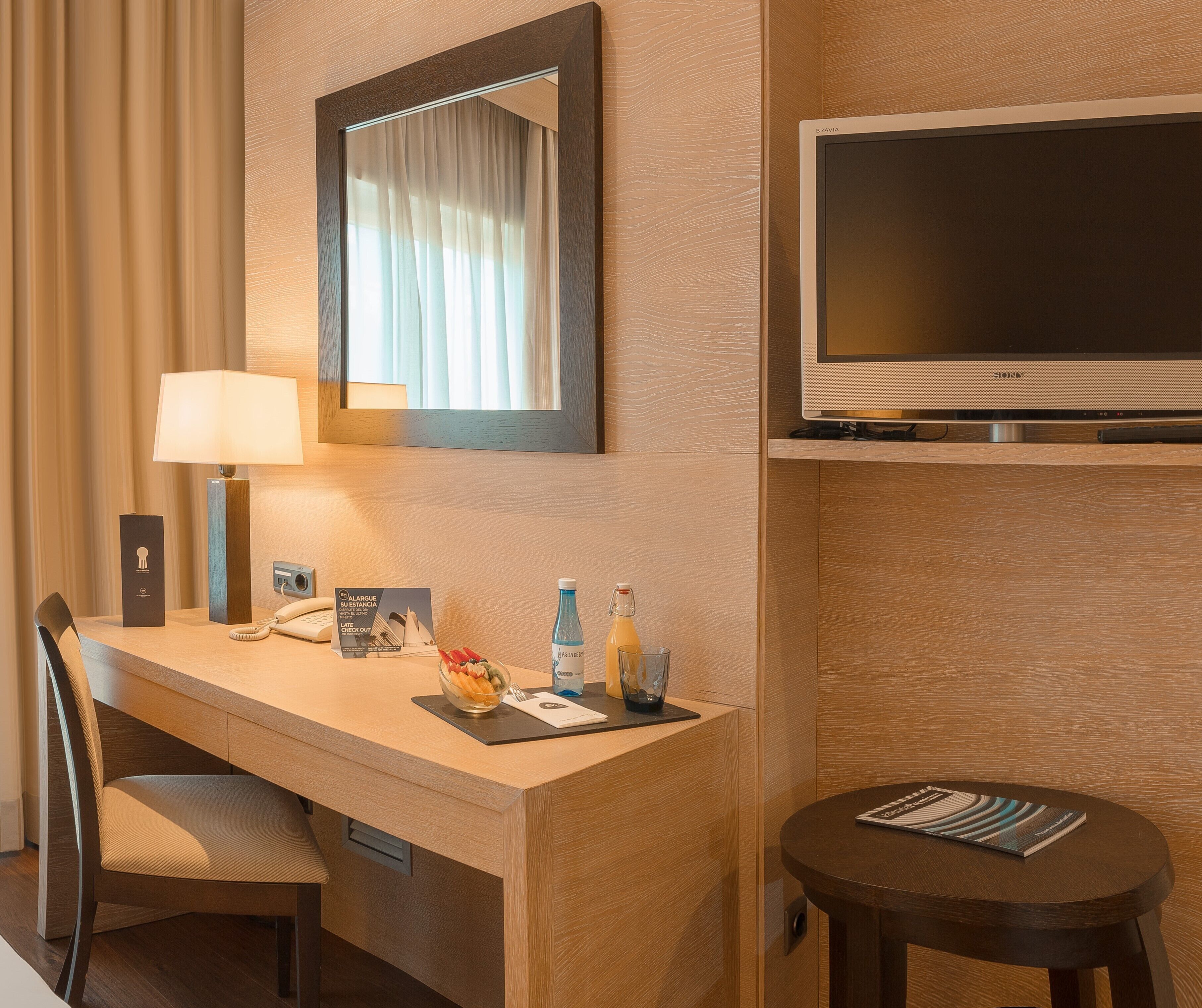 A hotel room with a desk, chair, lamp, television, and a small round table with a magazine.