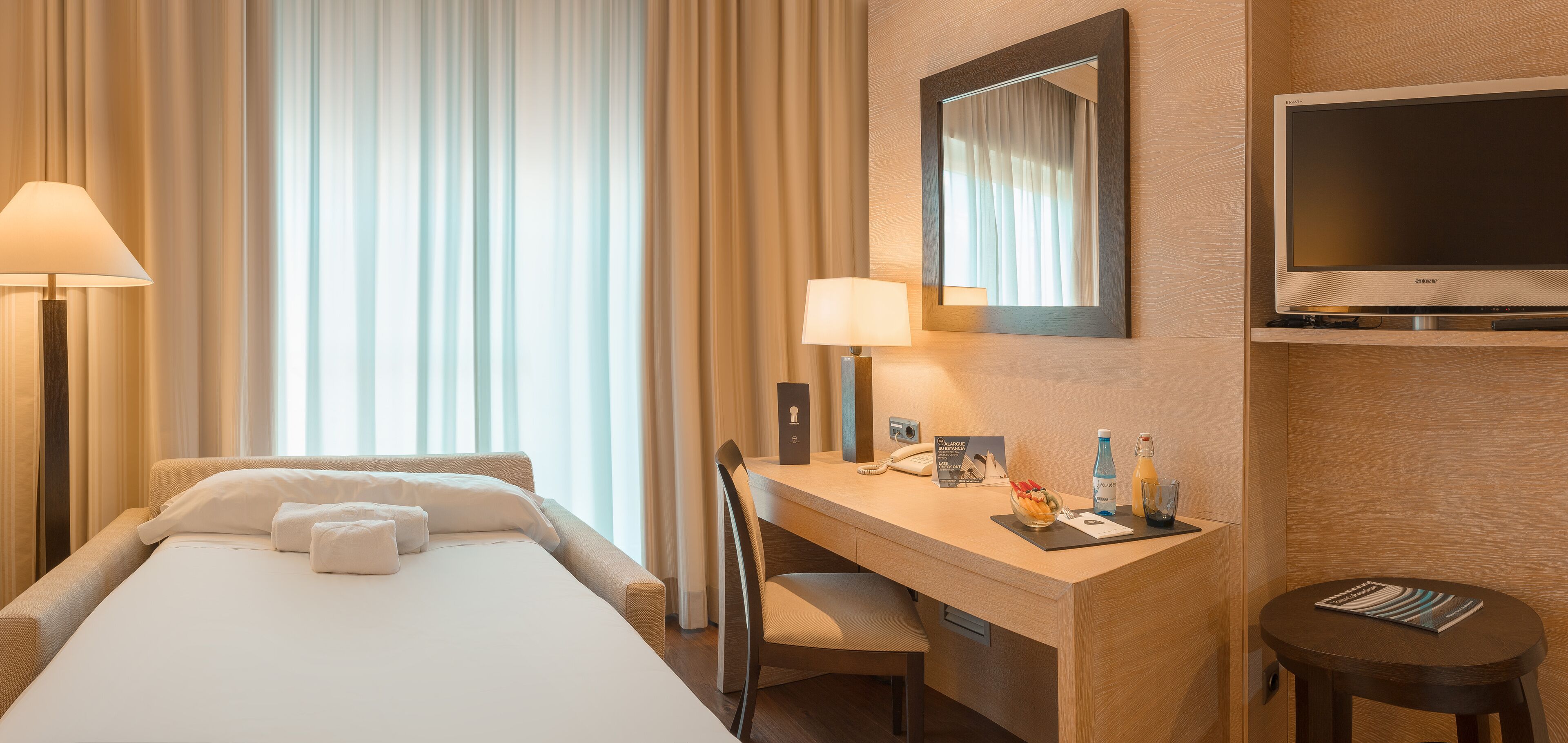 Quarto de hotel com cama, escrivaninha, cadeira, televisão e mesa de cabeceira com amenidades.