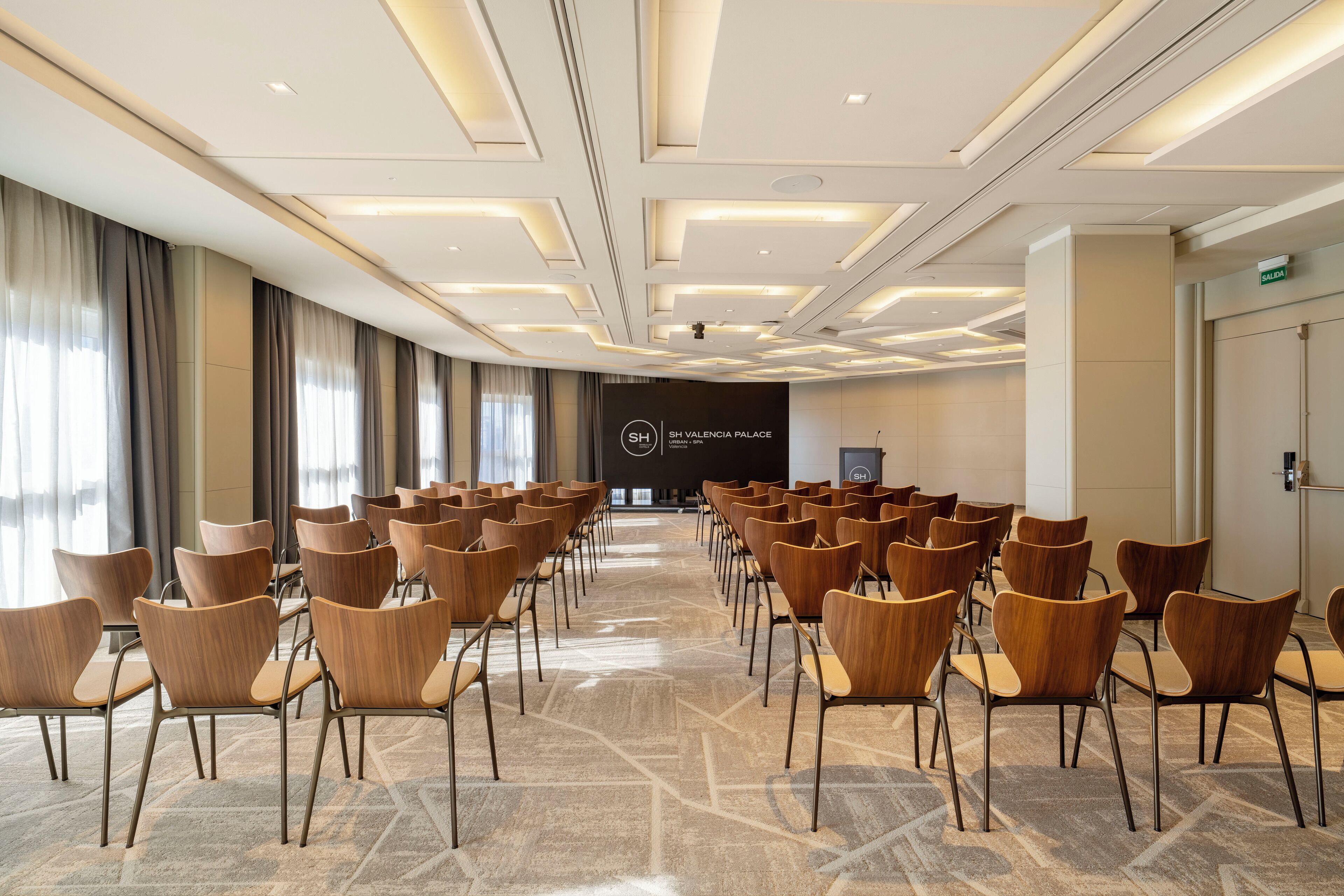 A conference room with rows of wooden chairs, a patterned carpet, and a modern ceiling design.
