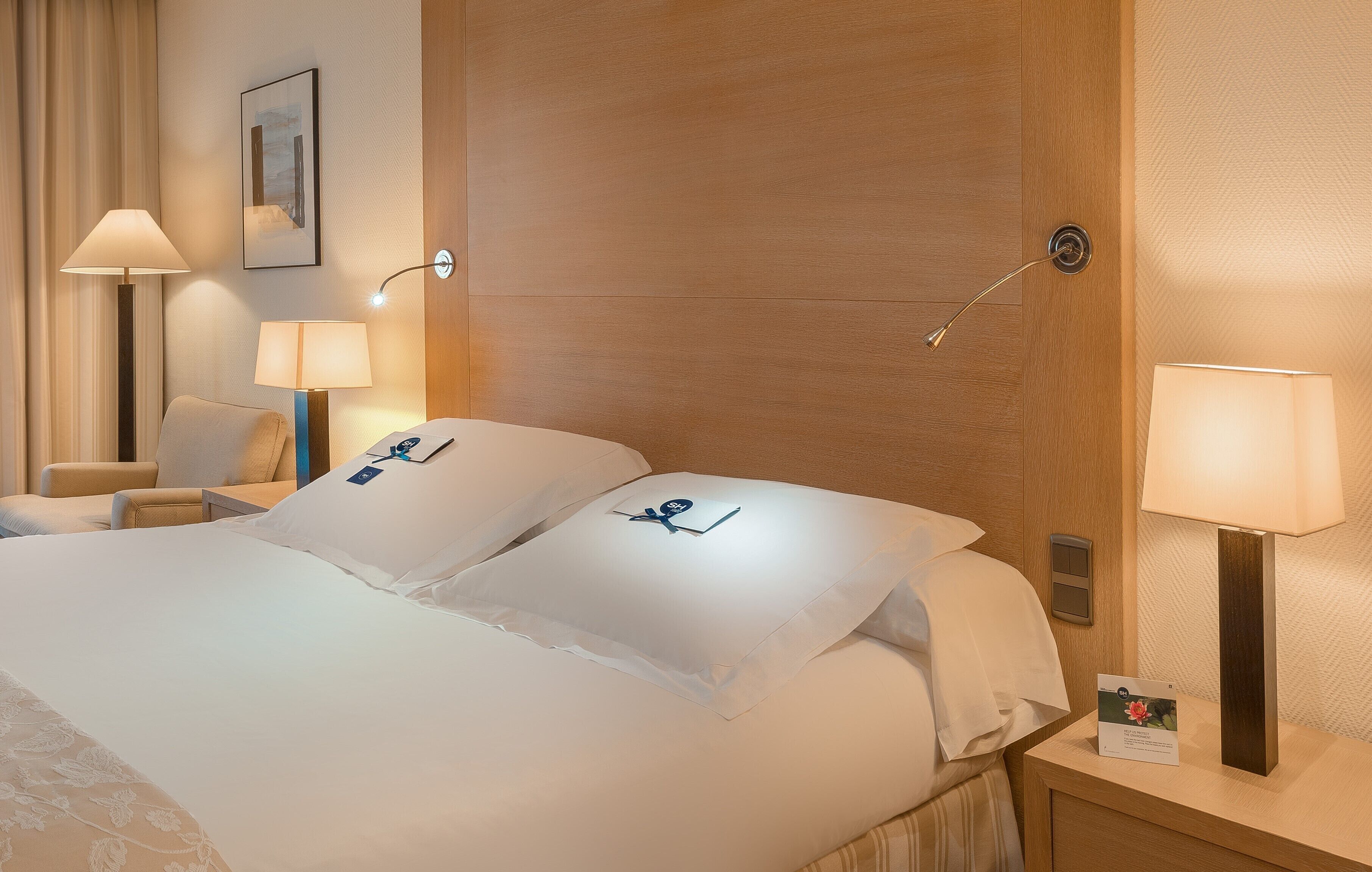 A neatly made bed with white linens, two bedside lamps, a wooden headboard, a small table with a lamp, and a framed picture on the wall.