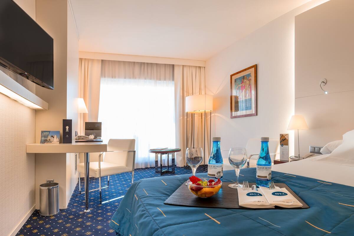 A hotel room with a bed, a desk, a chair, a TV, a window with curtains, and a fruit bowl on the bed.