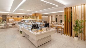 A modern hotel lobby with a central breakfast bar, seating areas, and a reception desk.