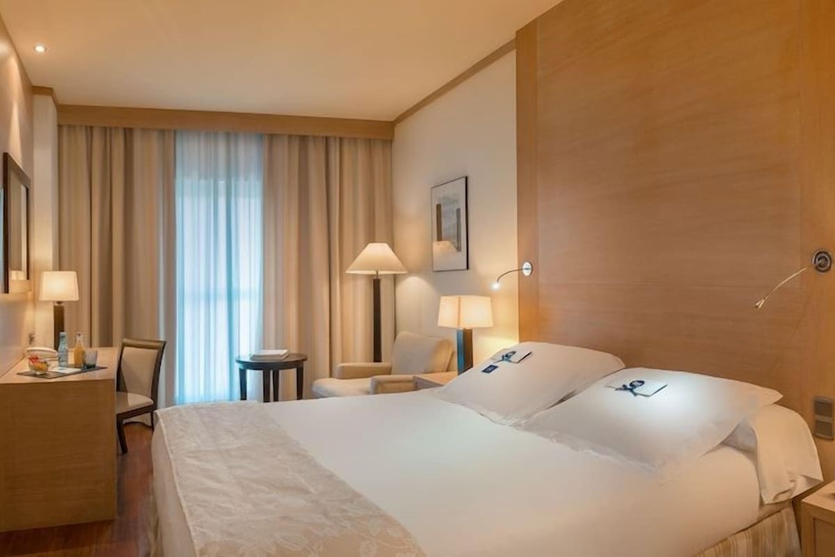 Habitación de hotel con cama, lámparas de noche, televisor, escritorio con silla y ventana con cortinas.