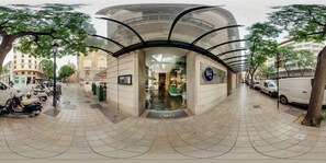 A modern building with a curved glass facade, a sign for 'SH', and a tiled pedestrian area with parked scooters. - SH Ingles Boutique Hotel (Valencia)