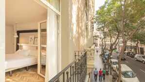 Une chambre d’hôtel avec un lit, une fenêtre et un balcon donnant sur une rue où sont garées des voitures et où circulent des piétons.