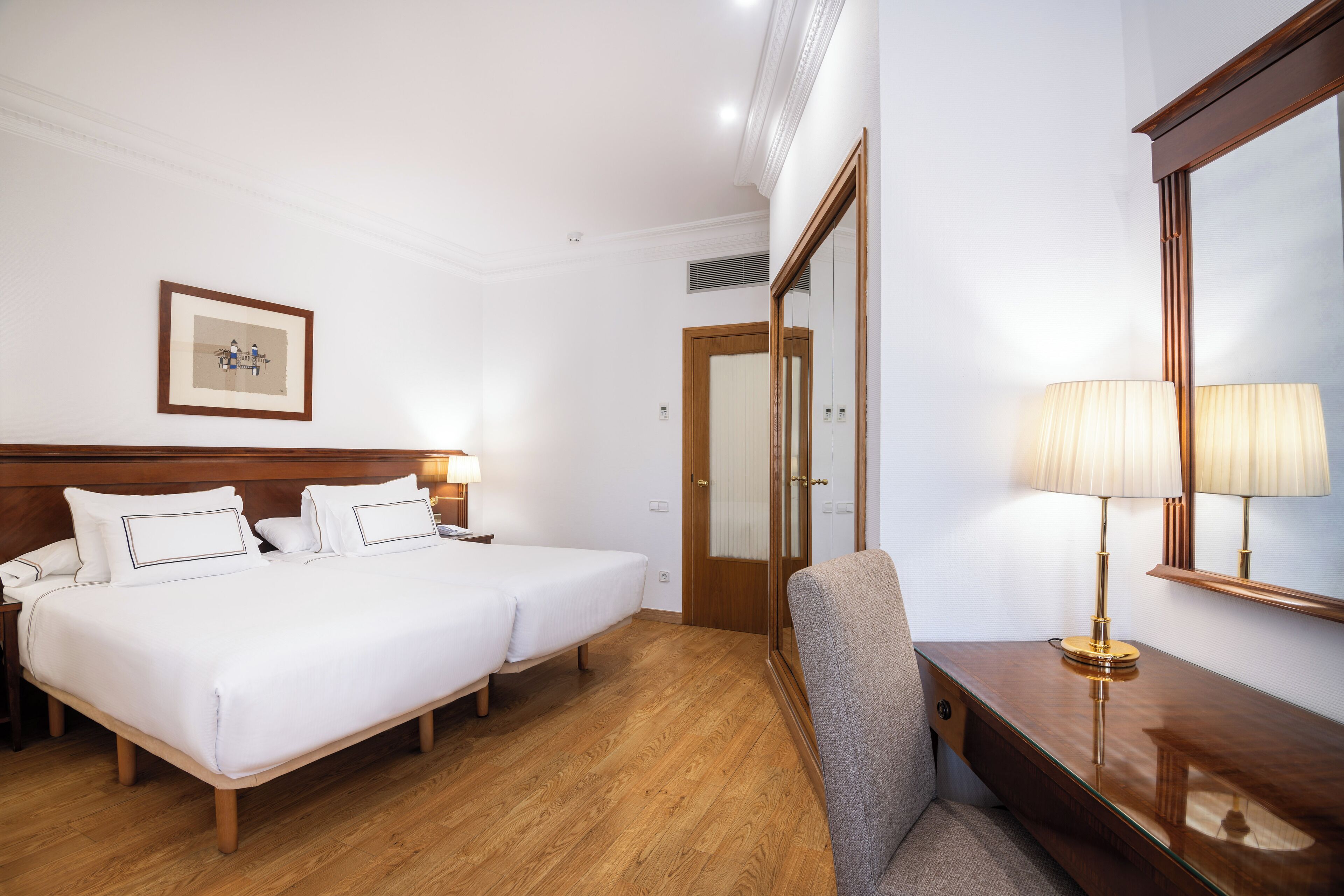 Une chambre d’hôtel avec deux lits, un bureau avec une lampe, une chaise, un tableau encadré au mur et un miroir au mur.