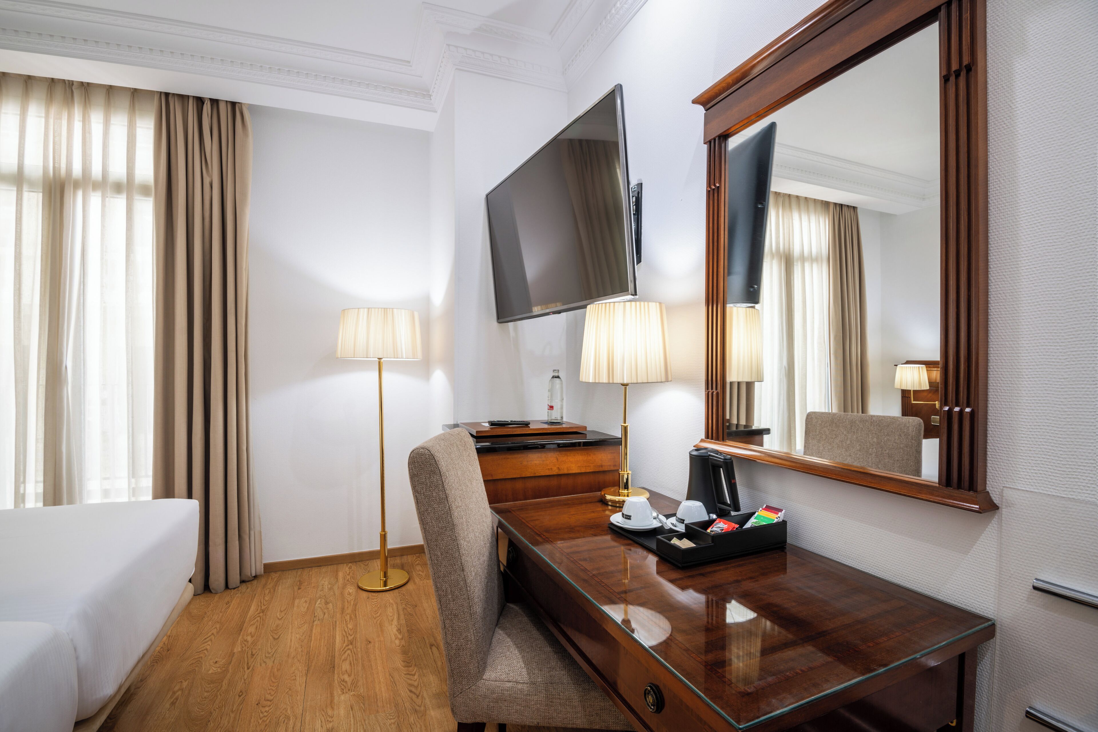 A hotel room with a bed, a desk with a flat-screen TV, a chair, a lamp, and a coffee maker.
