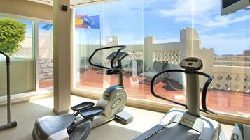 Un gimnasio con cintas de correr y equipo de ejercicio, ventanas amplias con vistas a edificios y una bandera, y un techo con un televisor montado.