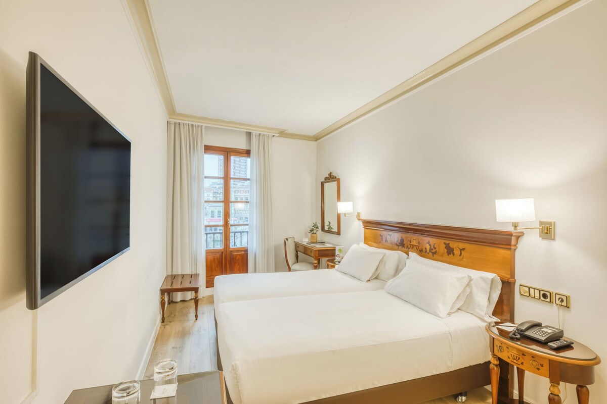 A hotel room with a large flat-screen TV, a wooden bed with white linens, a bedside table with a lamp, and a small desk with a chair.