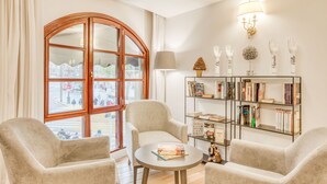 Una habitación acogedora con un amplio ventanal arqueado, una mesa redonda, dos sillones, una estantería y una lámpara fijada a la pared.