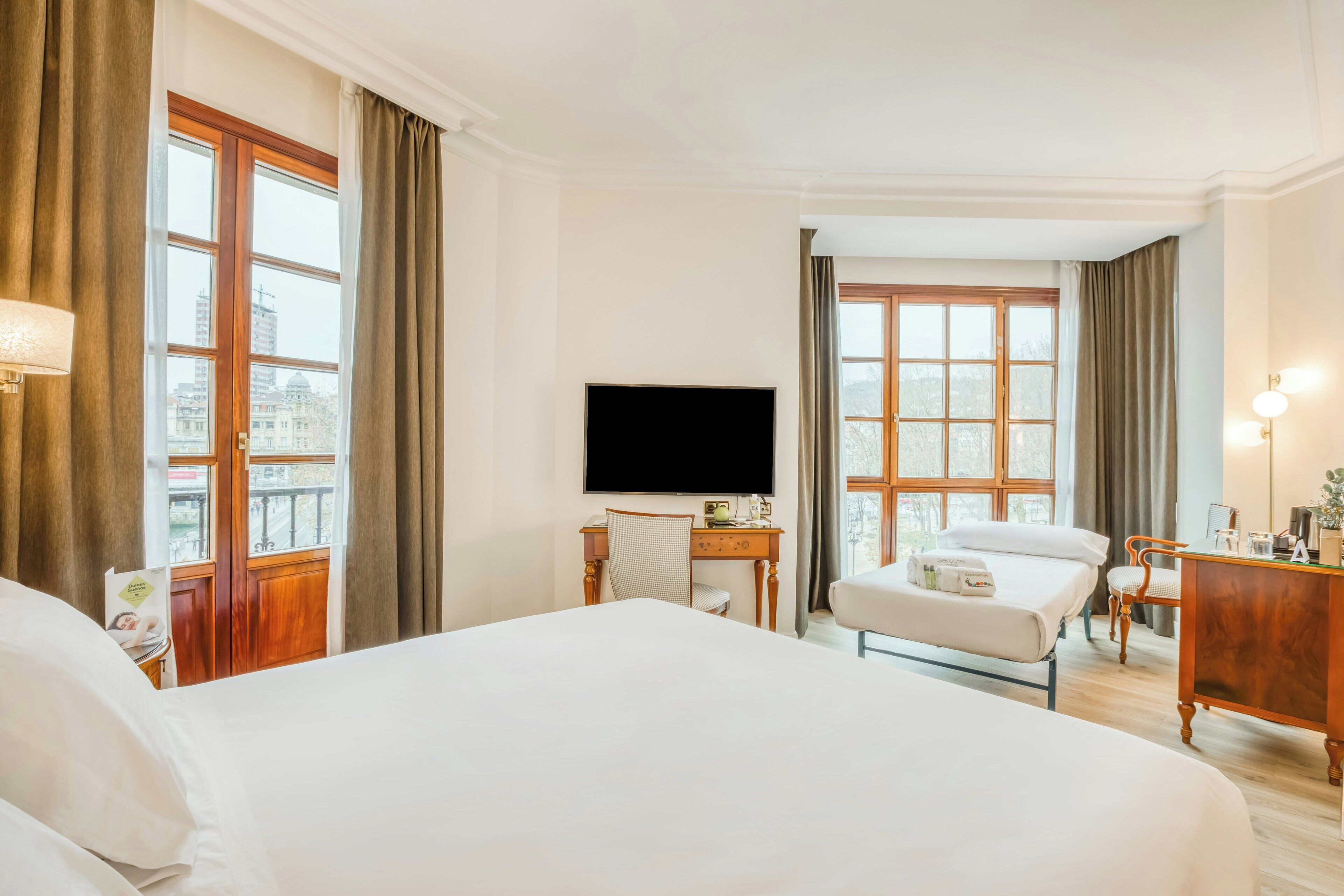 Habitación de hotel con una cama grande, un escritorio con televisor de pantalla plana, una silla y vistas de la ciudad a través de amplias ventanas.