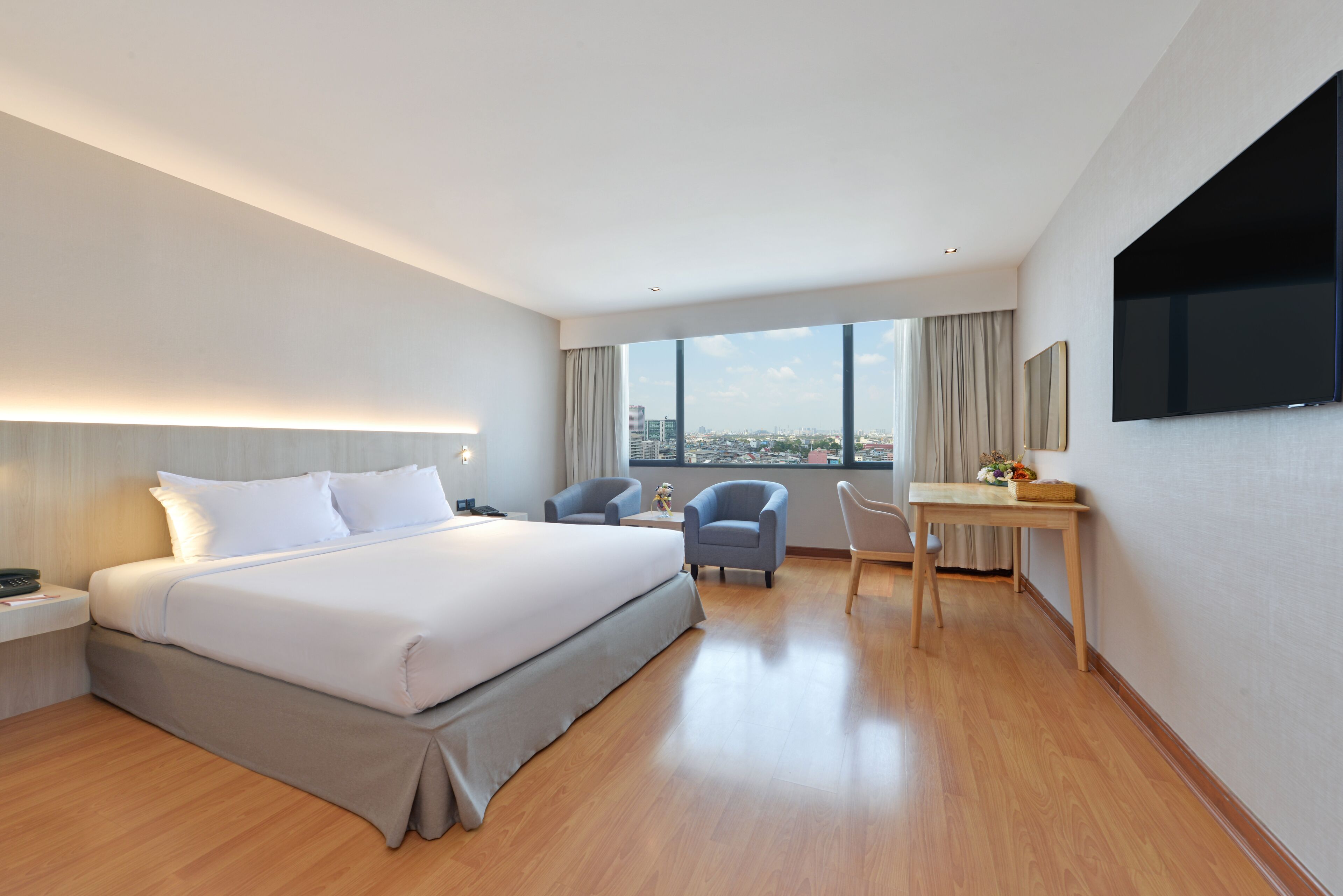 A modern hotel room with a large bed, a desk, two chairs, a flat-screen TV, and a city view.