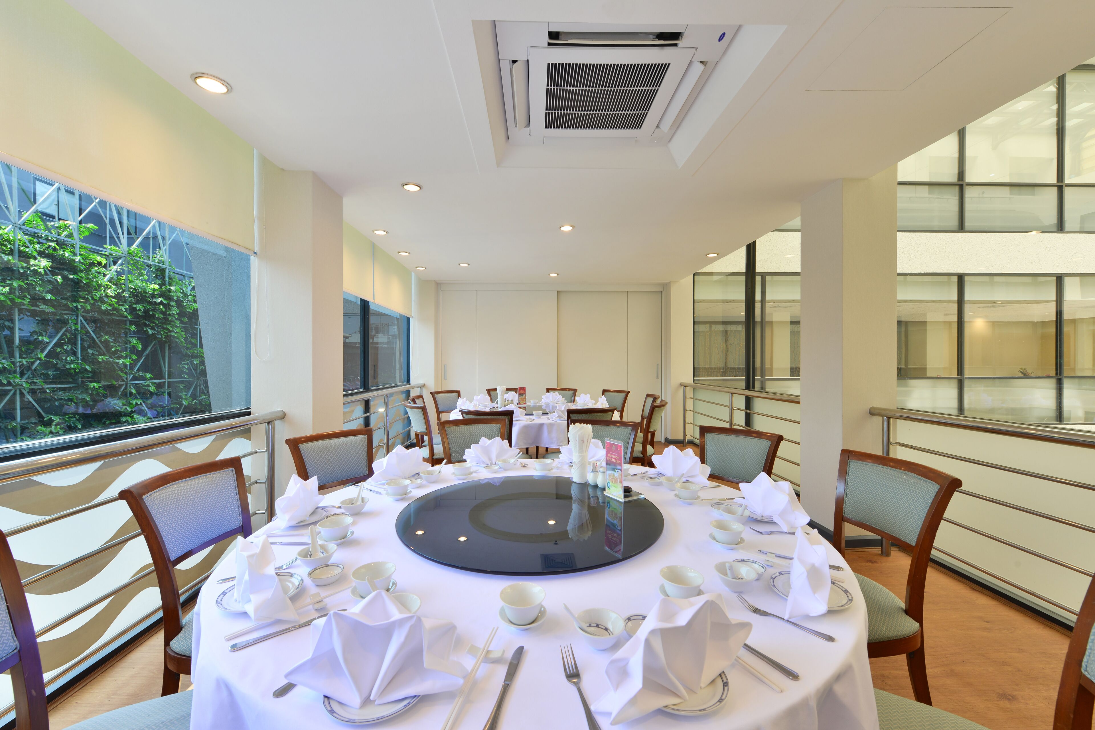 Un espace repas avec des tables rondes dressées pour le service, de grandes fenêtres et une vue sur la verdure environnante.