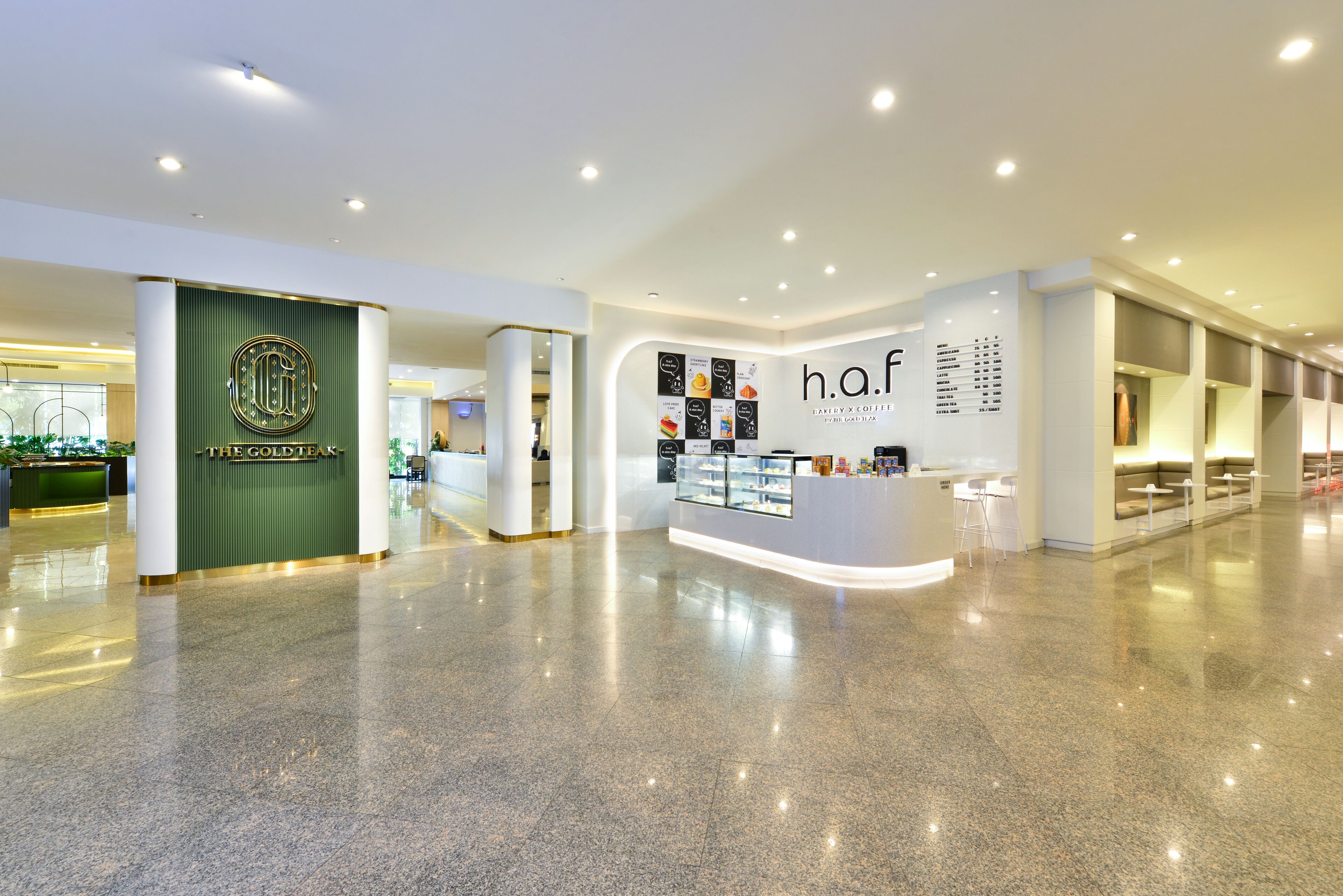 A modern hotel lobby with a reception desk, a coffee shop area, and a green wall with a logo.