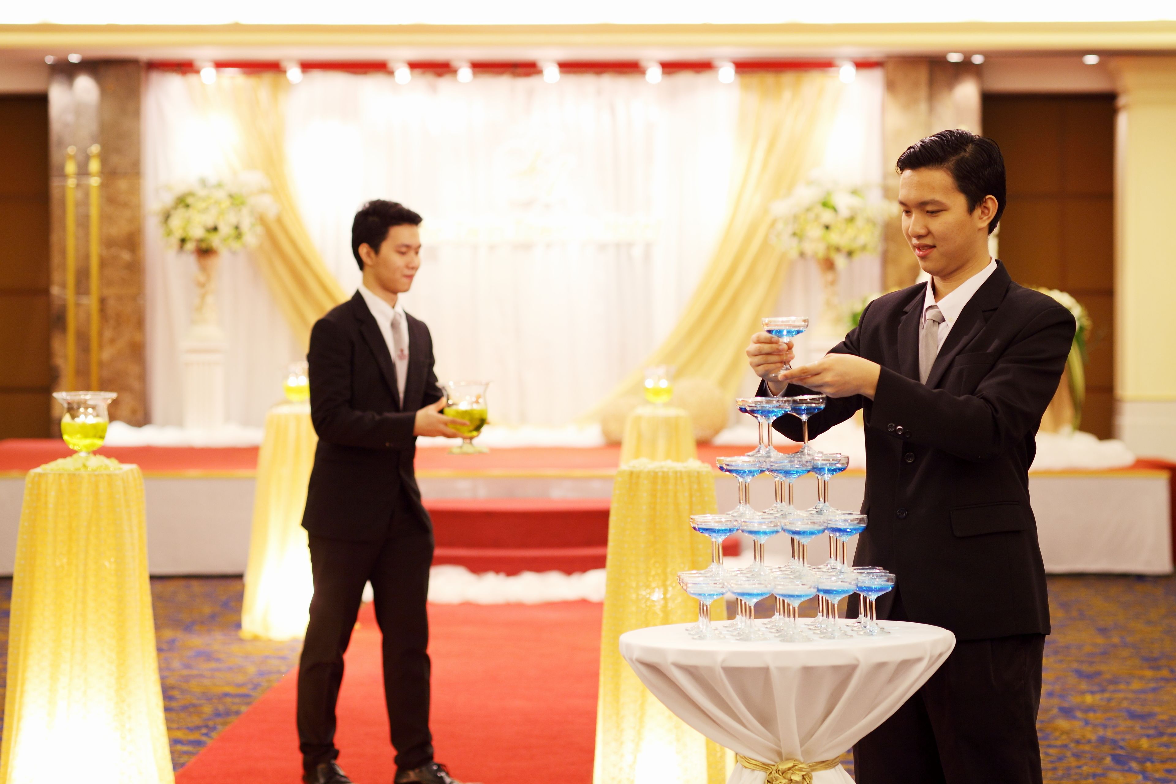 Dos hombres con traje en un evento formal, con una mesa de bebidas escalonada y una alfombra roja.