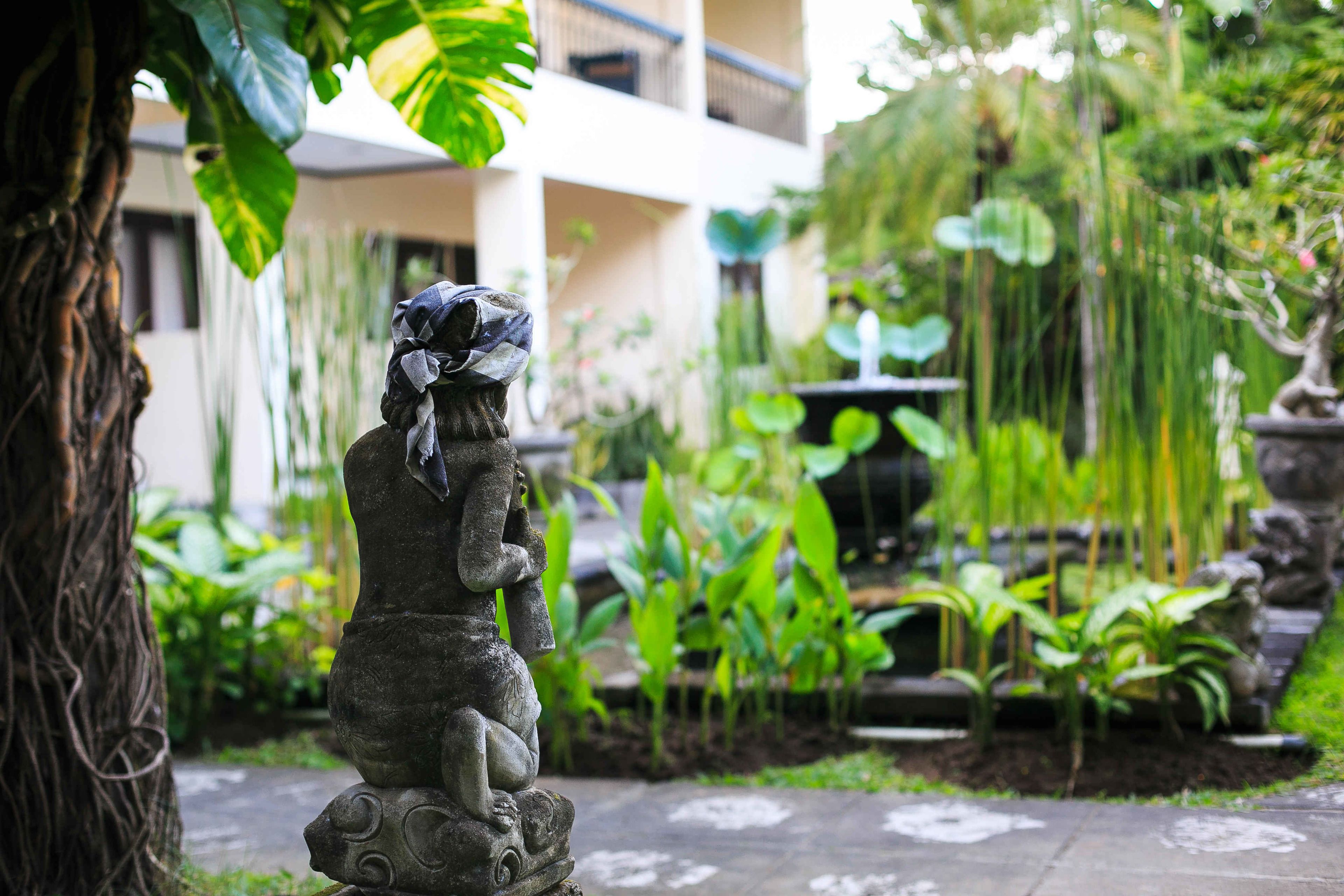 Un jardín con una estatua de piedra, abundante follaje y un edificio al fondo.