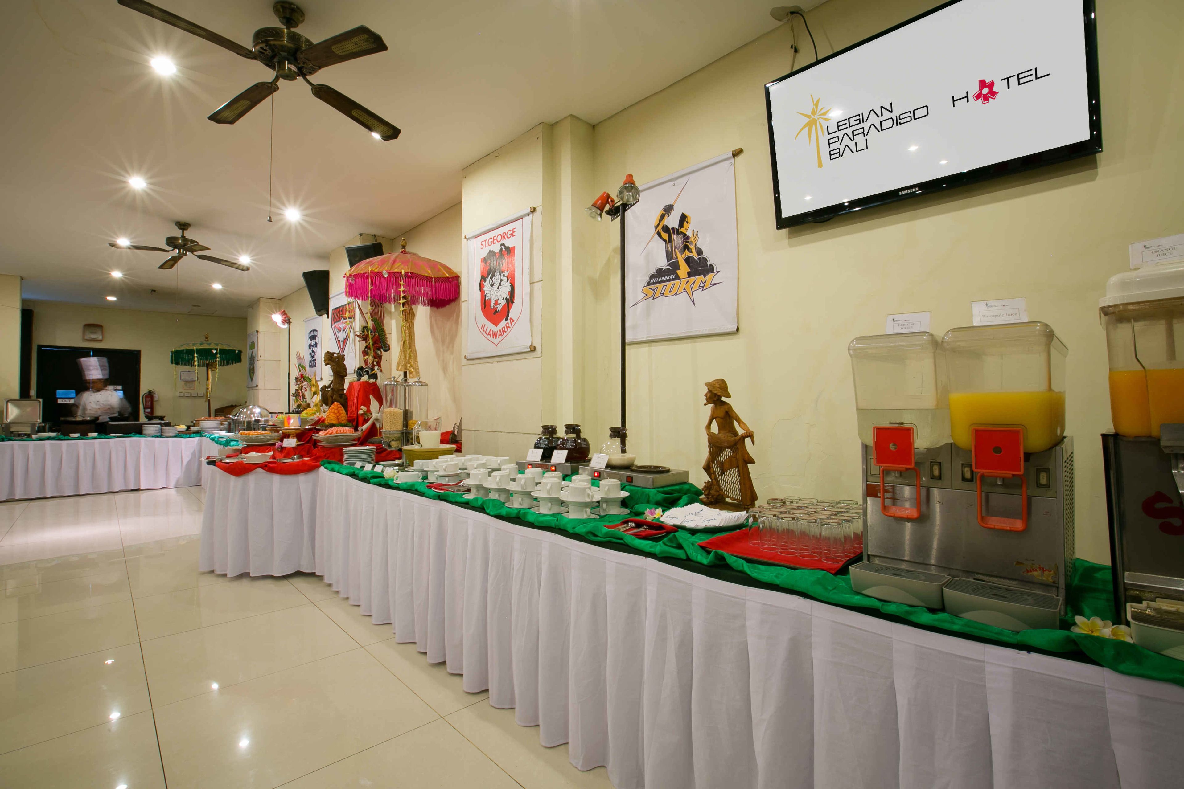 A buffet setup with various food items and a beverage station, a ceiling fan, and a television screen displaying hotel information.