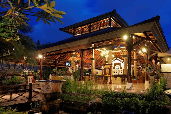 A traditional wooden building with a tiled roof, surrounded by lush greenery and a water feature in the foreground. - Legian Paradiso Hotel (Legian)