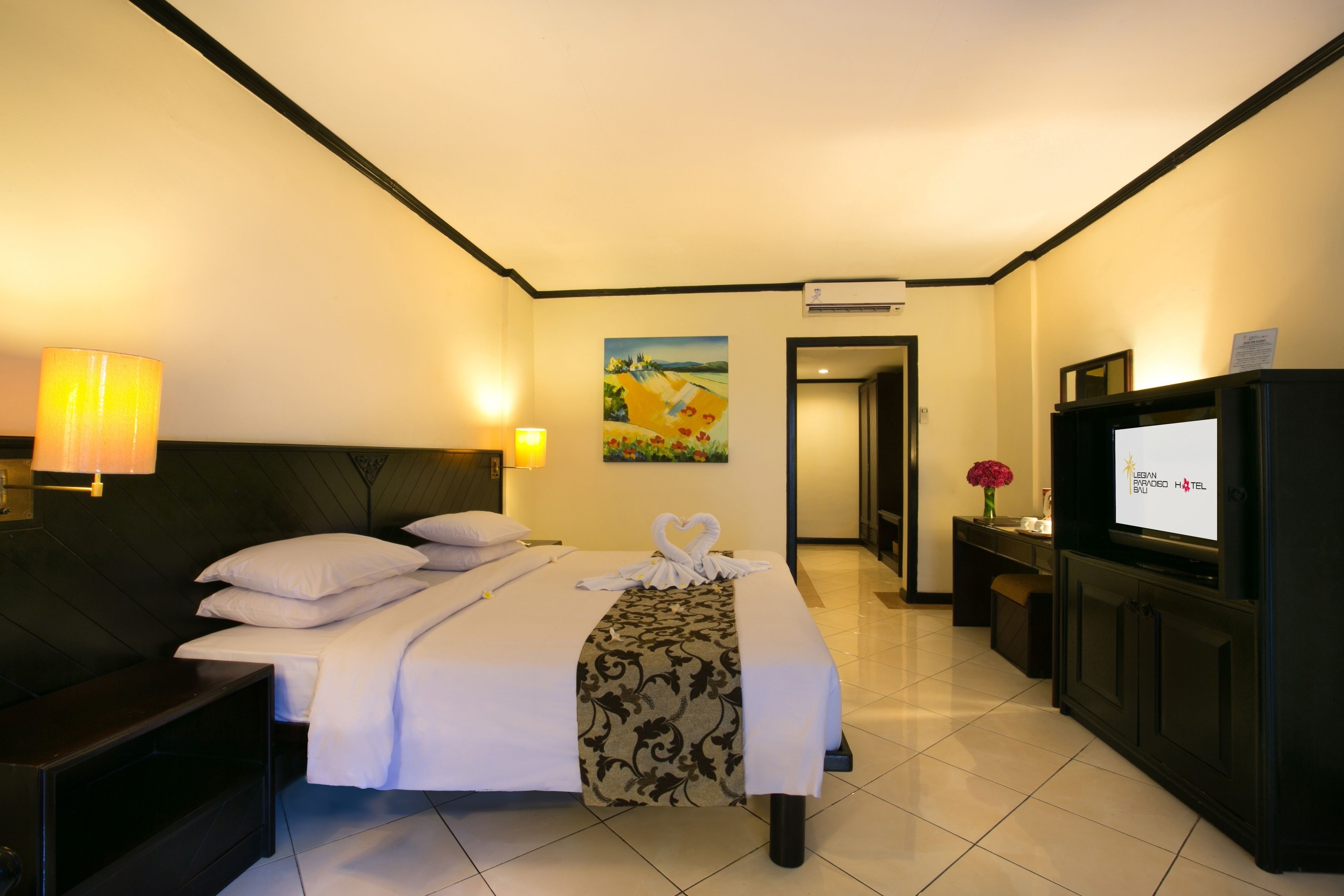 Una habitación de hotel con cama, mesita de noche, lámpara, televisor y una obra de arte en la pared.