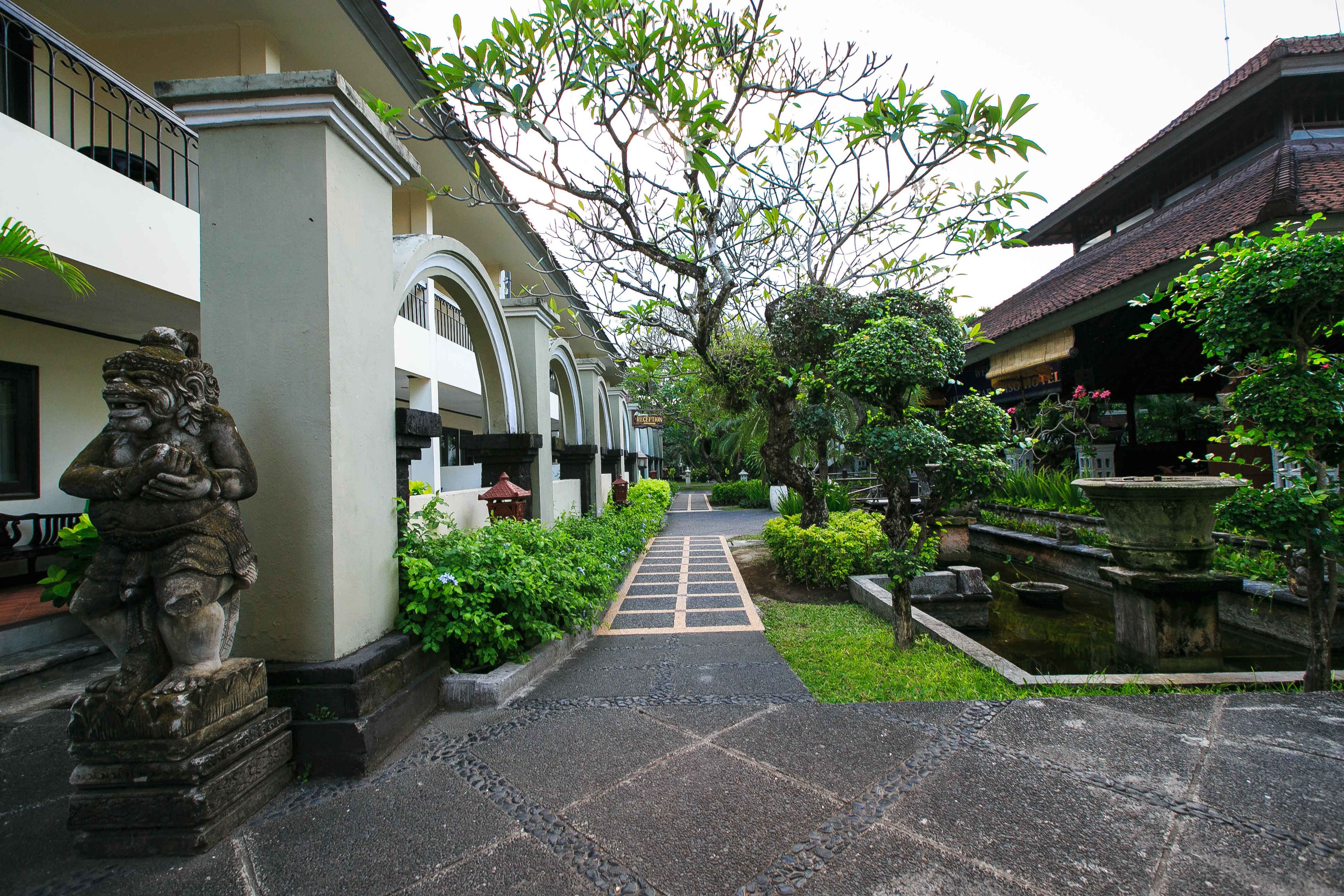 A pathway leading to a building with a statue on the left, surrounded by greenery and a small pond.