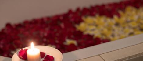 A lit candle on a plate with rose petals, surrounded by a bed of red and yellow flowers.