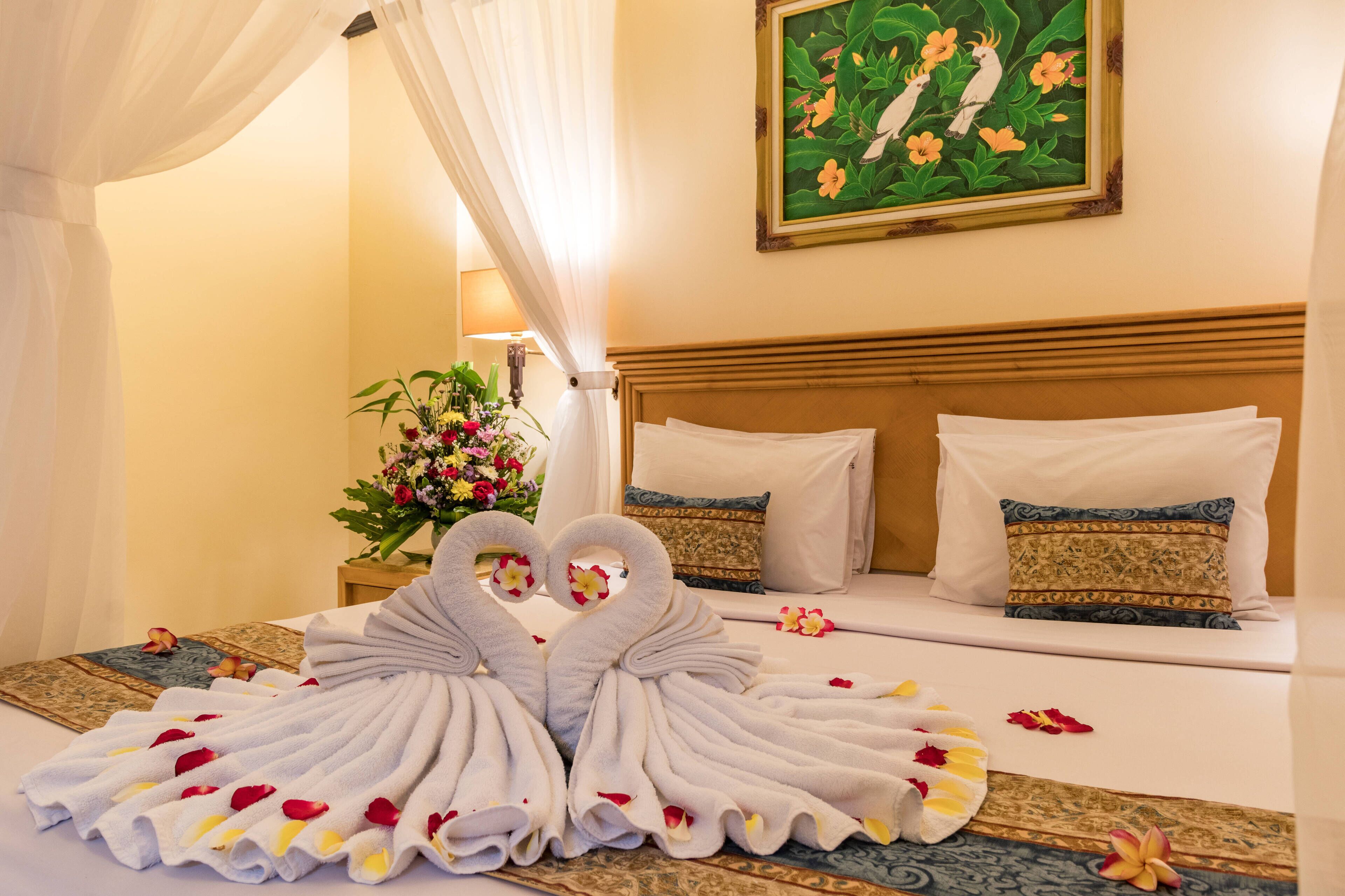 Quarto de hotel com cama, duas almofadas, um buquê de flores e um quadro na parede.