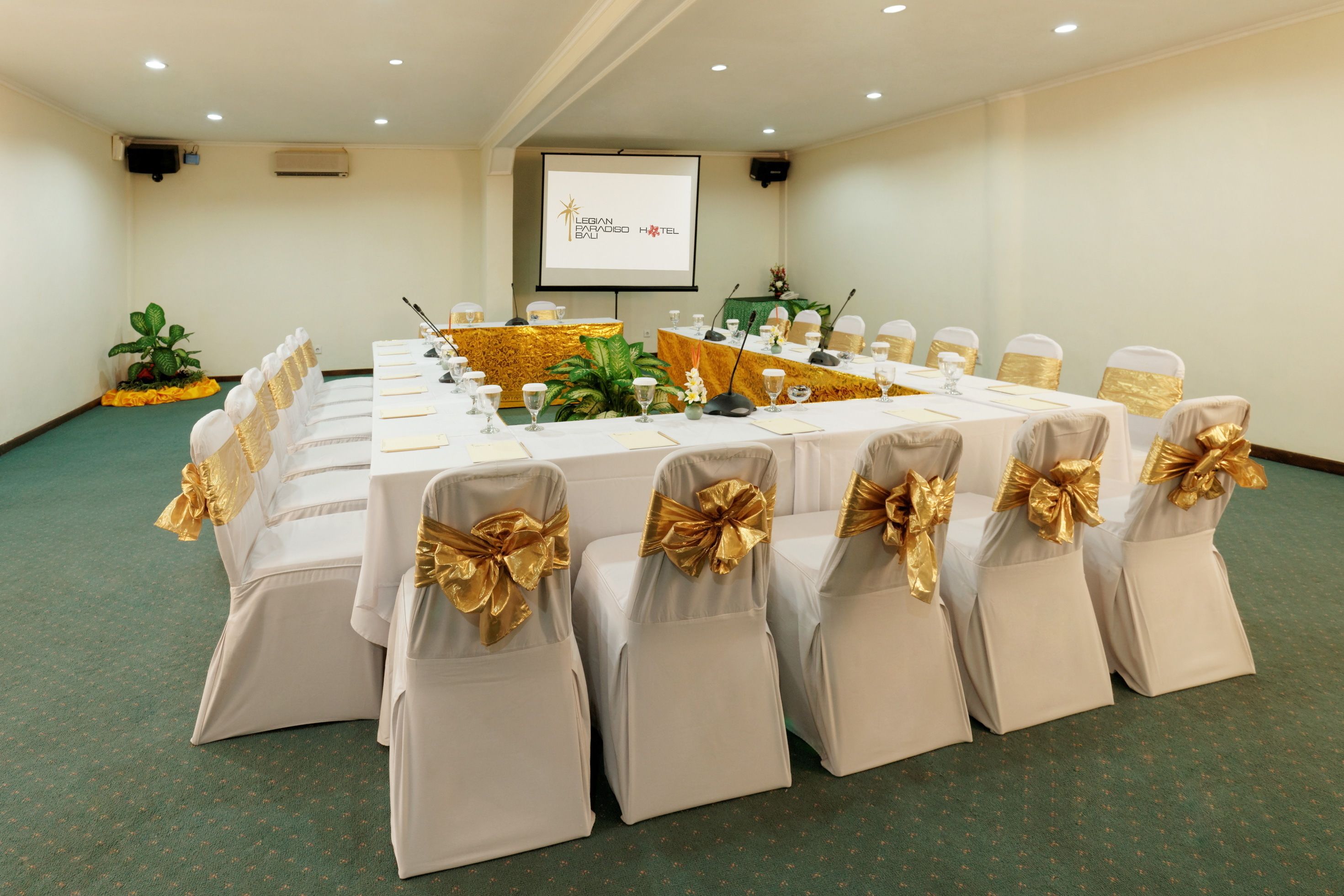A conference room with a long table, white chairs with gold sashes, and a projector screen displaying "LEBAN ESPAÇO HOTEL".