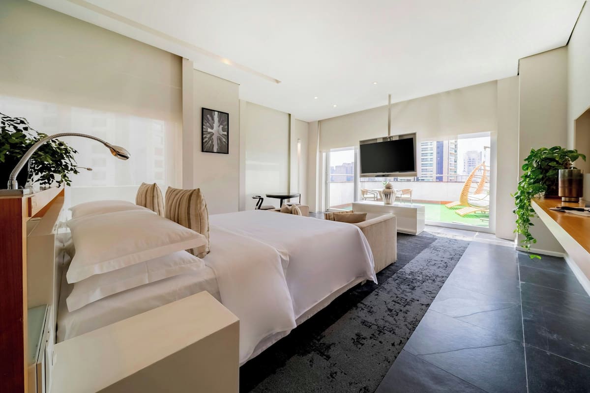 A modern hotel room with a large bed, a desk, a TV, and a balcony with a view of the city.