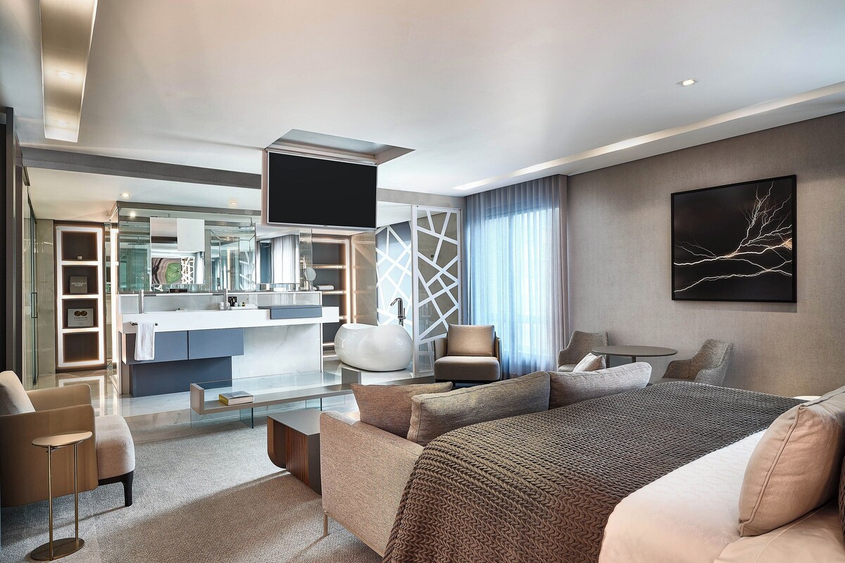 Une chambre d’hôtel moderne dotée d’un grand lit, d’un canapé, d’une télévision et d’une salle de bains avec un grand miroir, ainsi que d’un coin repas.