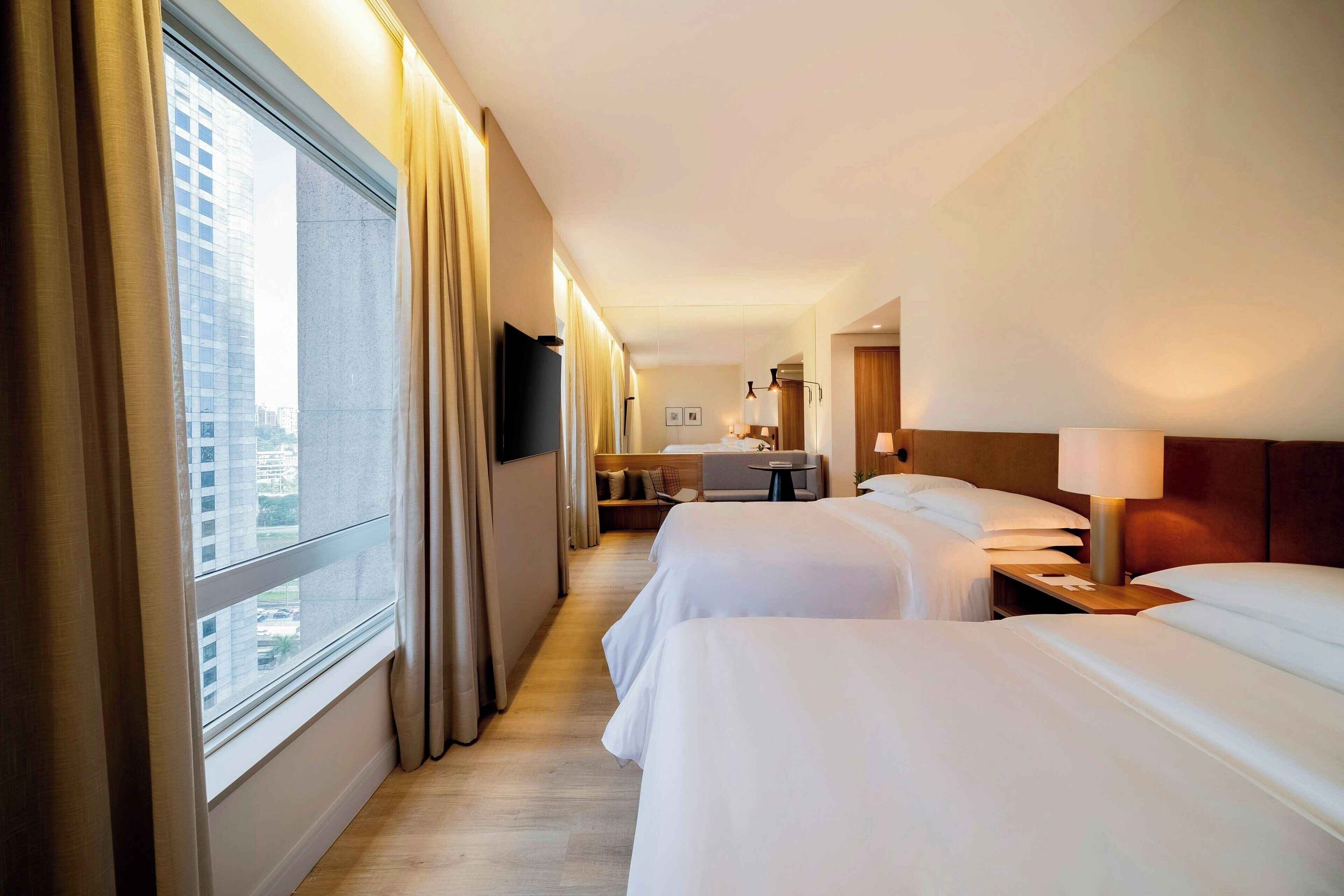 Ein Hotelzimmer mit zwei Betten, einem Schreibtisch, einem Stuhl, einem Fernseher und einem großen Fenster mit Vorhängen.
