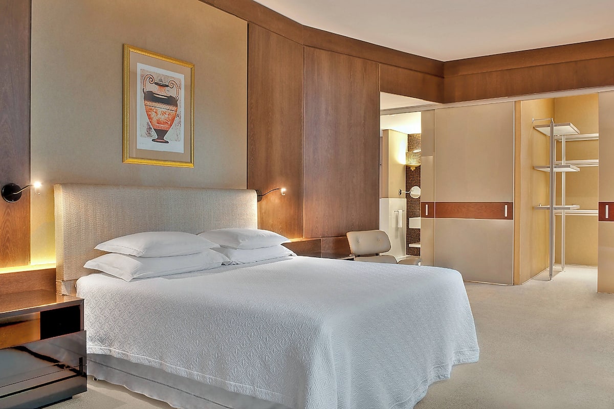 A hotel room with a large bed, a bedside table, a nightstand, a framed picture on the wall, and a walk-in closet with shelves.