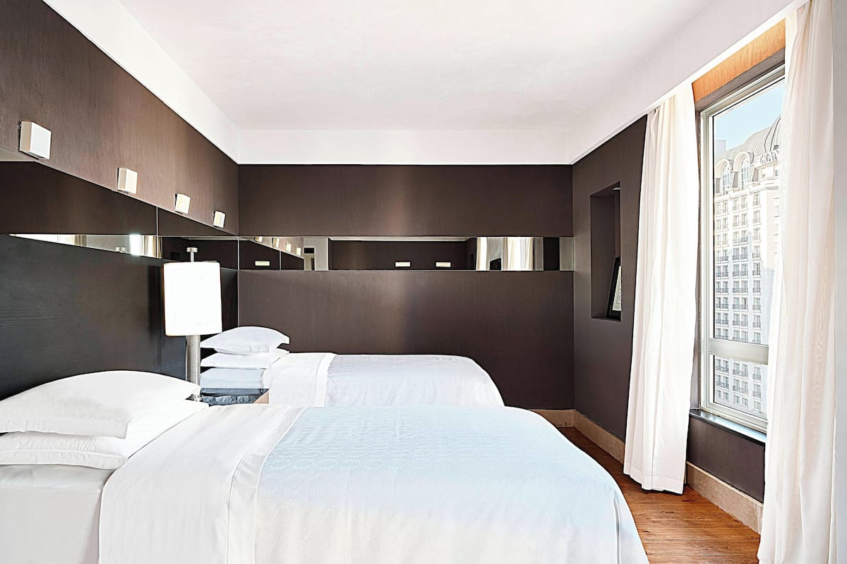 A modern hotel room with two beds, a large window with curtains, and a city view.