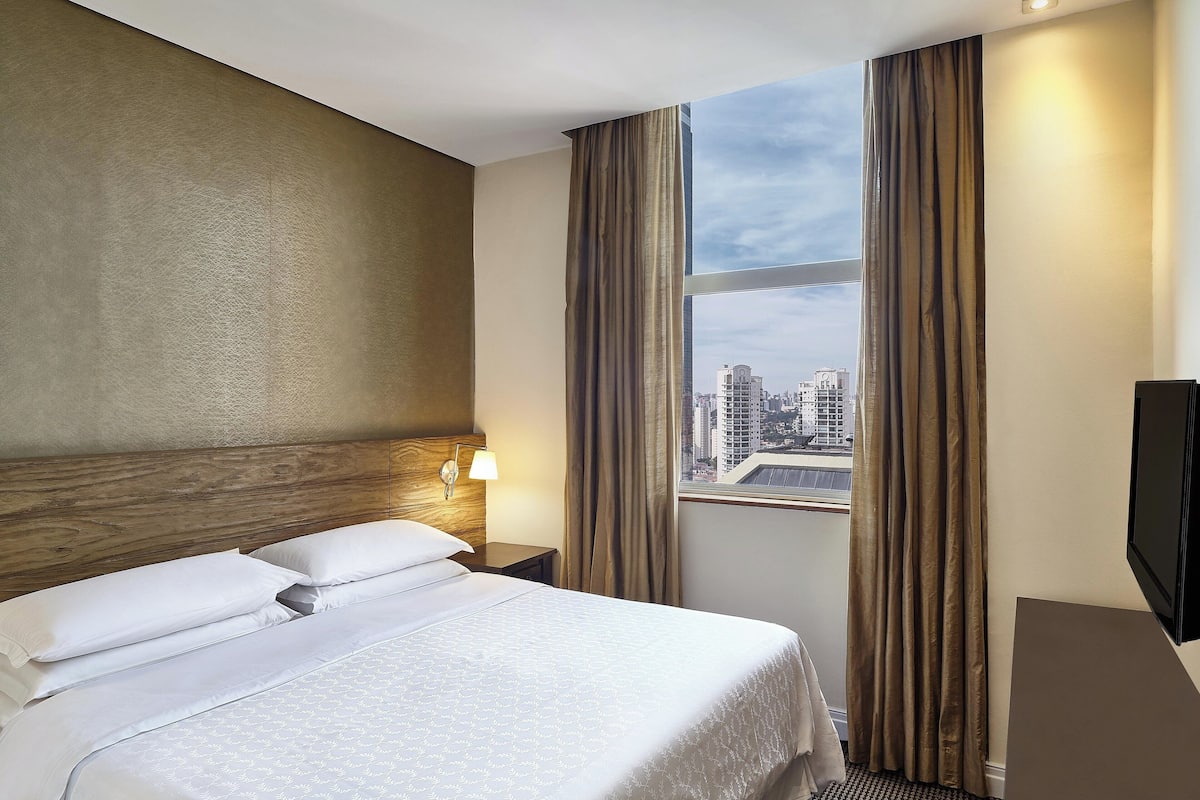 A hotel room with a large bed, a wooden headboard, a nightstand with a lamp, a TV, and a window with curtains.