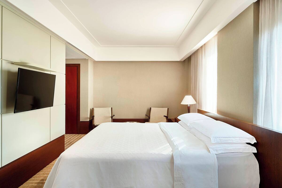 A hotel room with a large bed, a flat-screen TV mounted on the wall, two bedside tables with lamps, and a window with curtains.