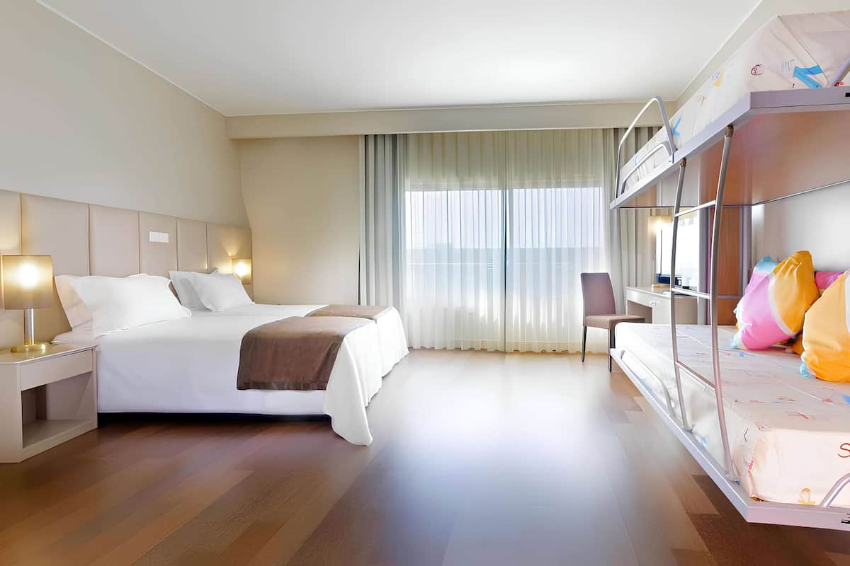A hotel room with a bed, bedside tables, a chair, a desk, and a bunk bed.