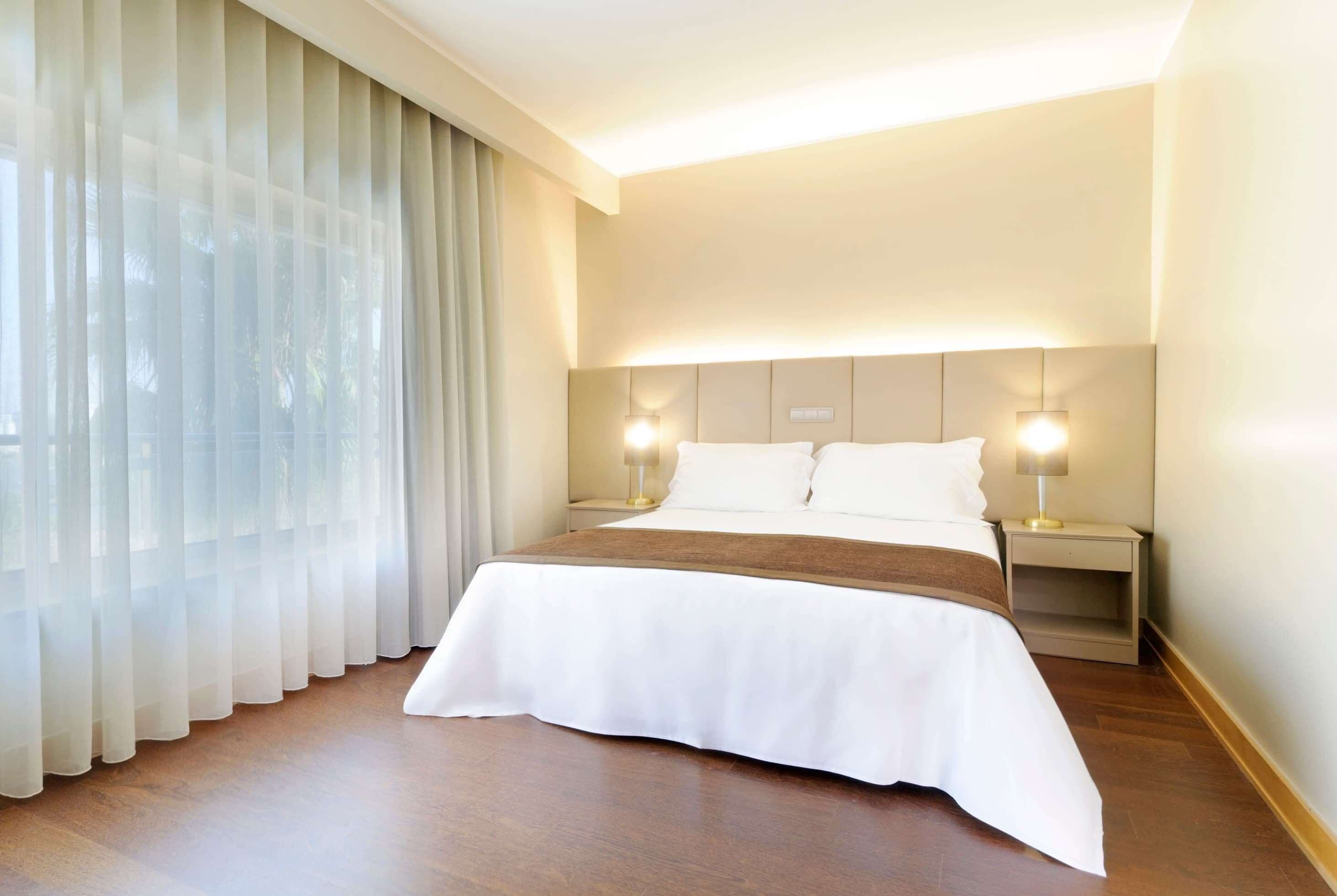 A hotel room with a large bed, two bedside lamps, a headboard, and sheer curtains.