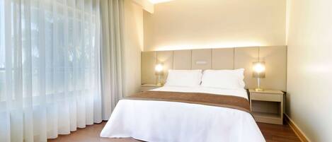 A hotel room with a large bed, two bedside lamps, a headboard, and sheer curtains.