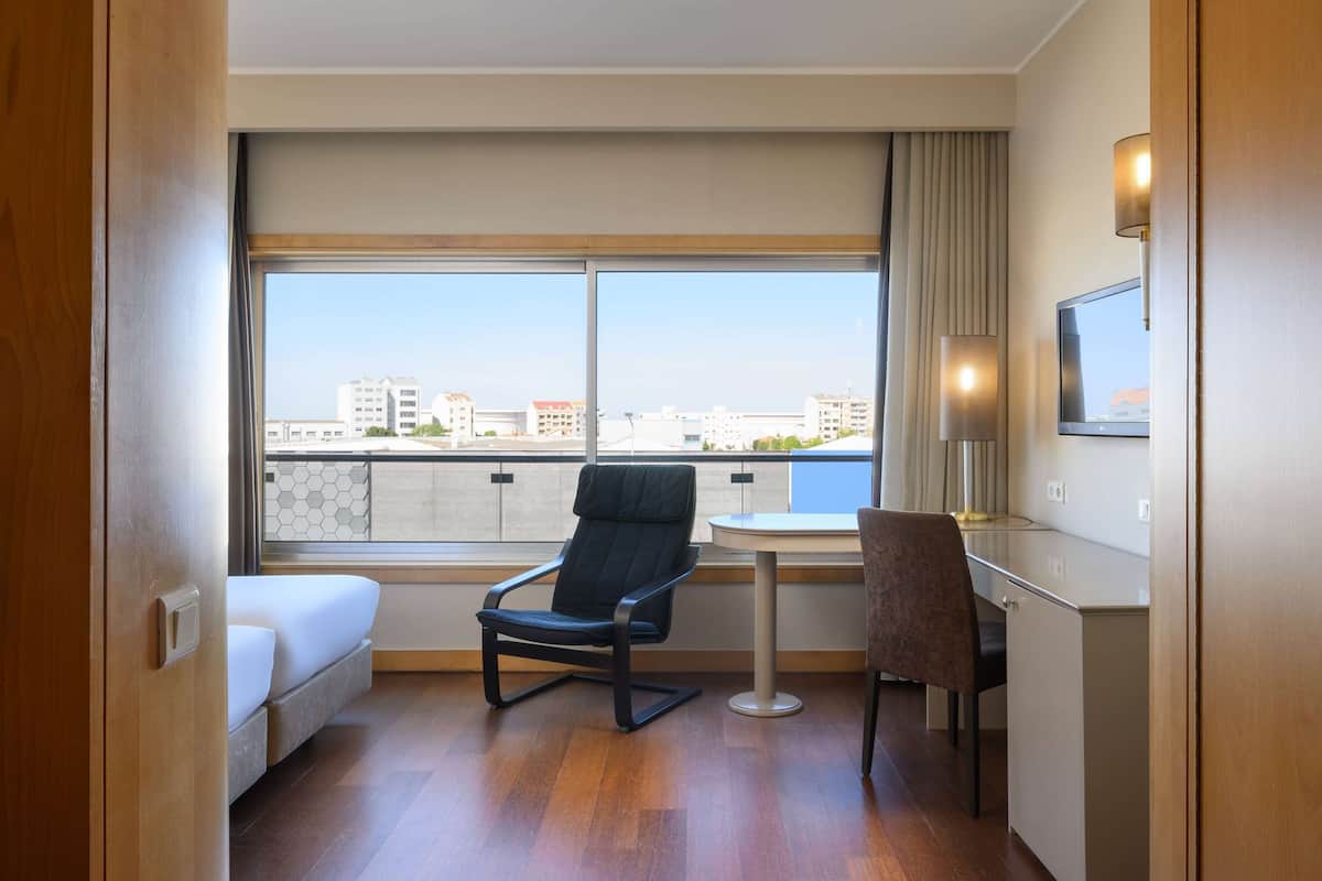 A hotel room with a bed, a desk, a chair, a TV, and a large window with a city view.