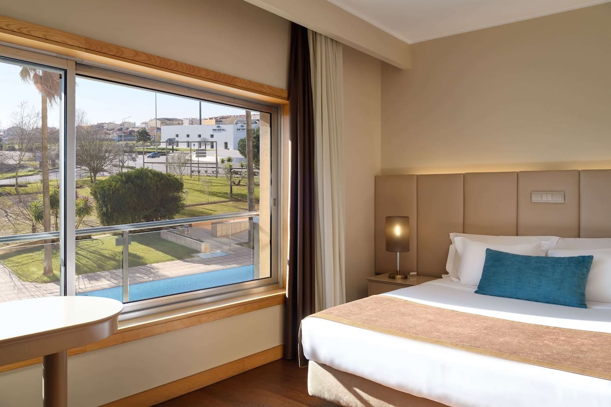 A hotel room with a large window, a bed with white bedding and a blue pillow, a bedside table, and a lamp.
