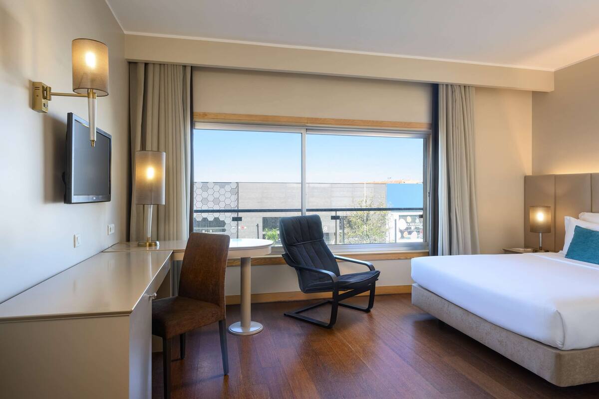 A hotel room with a bed, a desk, a chair, a TV, and a large window with curtains.