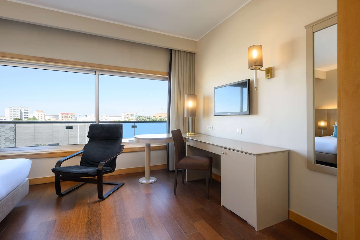 A hotel room with a bed, a desk, a chair, a TV, and a large window offering a city view.