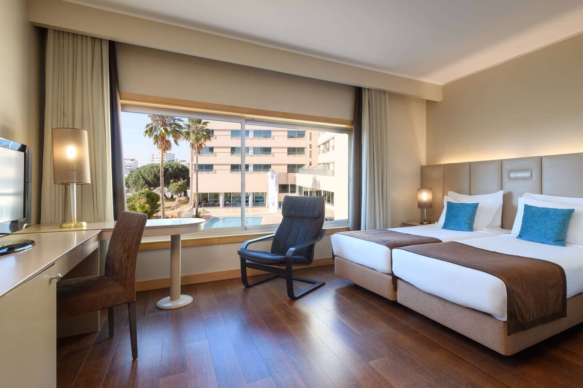 A hotel room with a desk, chair, two beds, and a large window with a view of buildings and palm trees.