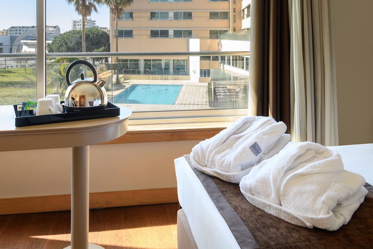 A hotel room with a view of a swimming pool, a table with a kettle and tea bags, and a bed with folded towels.
