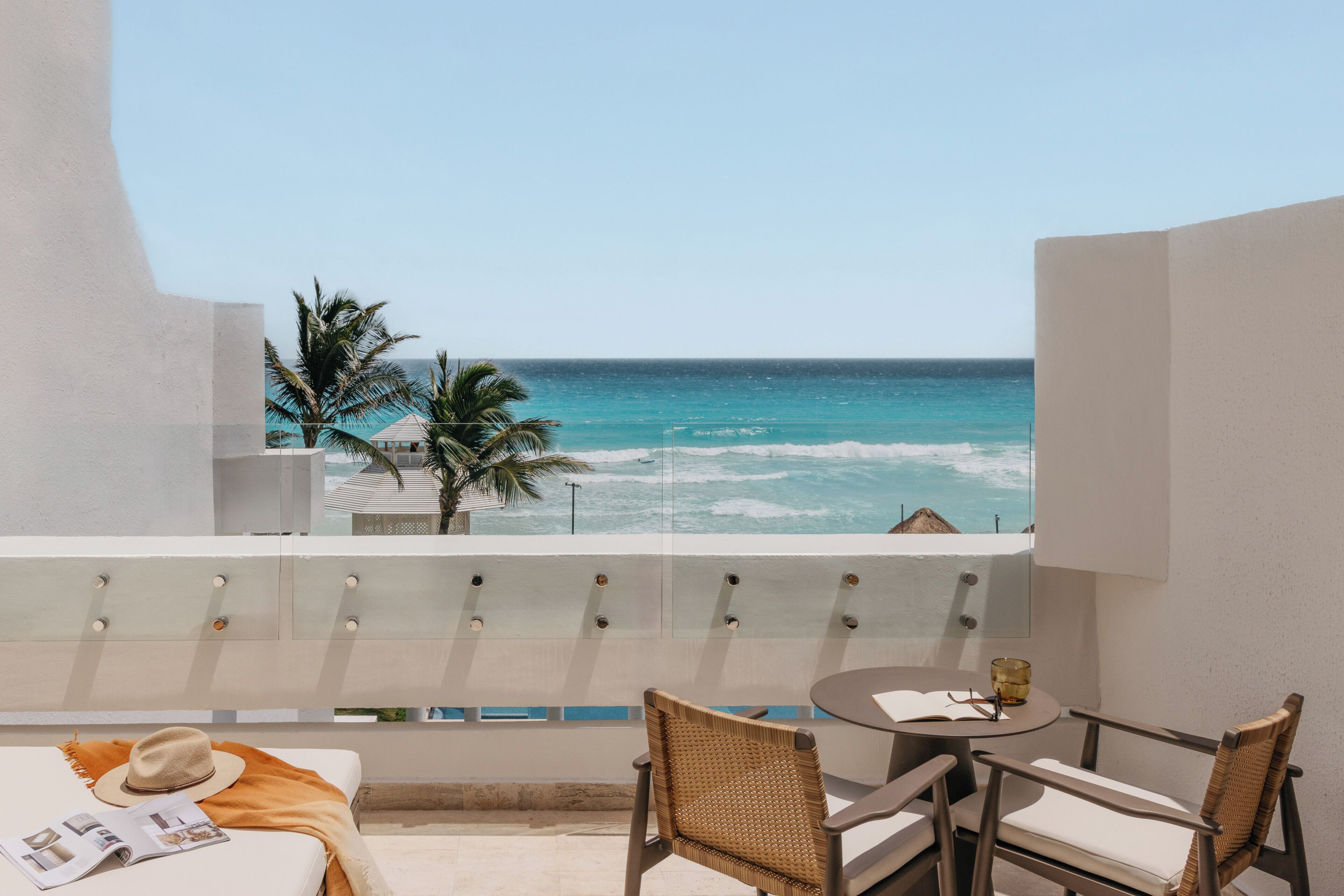 A balcony with a view of the ocean, a table with chairs, and a bed with a magazine and hat.