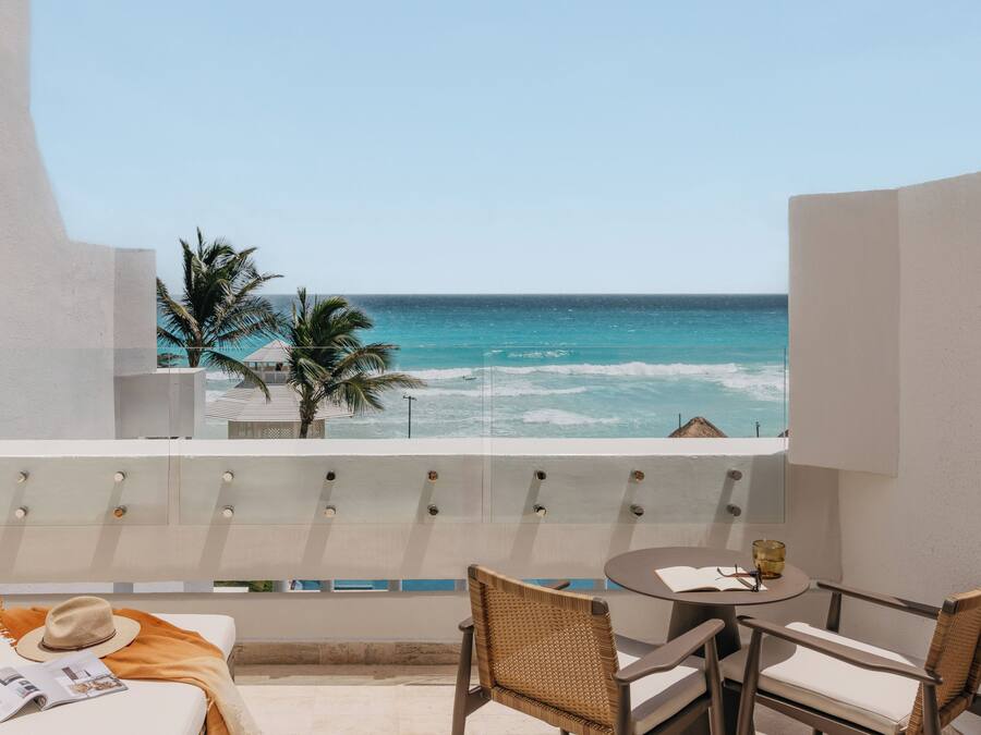 A balcony with a view of the ocean, a table with chairs, and a bed with a magazine and hat.